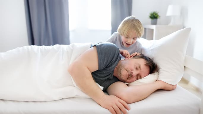 A middle-aged man is asleep in bed with a young child waking him up